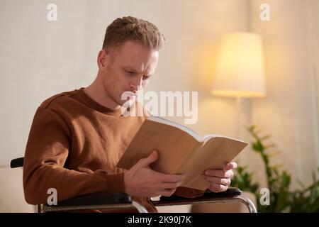 Ein junger Mann, der im Rollstuhl sitzt und ein Buch über seine Krankheit liest, runzelte die Stirn Stockfoto