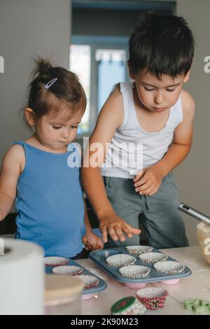 Zwei kleine kaukasische Kinder füllen den Teig in Cupcake-Silikonformen in der Küche. Konzept des Süßwarenkochens. Muffins machen. Roher Teig Stockfoto