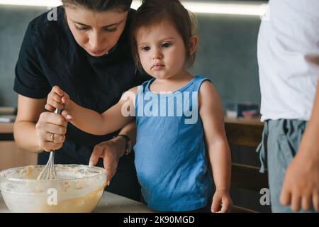 Kleines Mädchen mit ihrer Mutter, die Zutaten in einer großen Schüssel mischte, um Teig für Muffins zu machen. Kinder kochen mit den Eltern. Backen, Kuchendekoration, glücklich Stockfoto