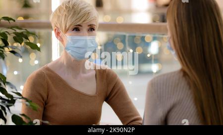 Zwei Freundinnen, Mutter mit Tochter im Teenageralter, die im Café spricht, tragen medizinische Gesichtsschutzmasken. Erwachsene 40s Frau Berater beraten Client in Stockfoto