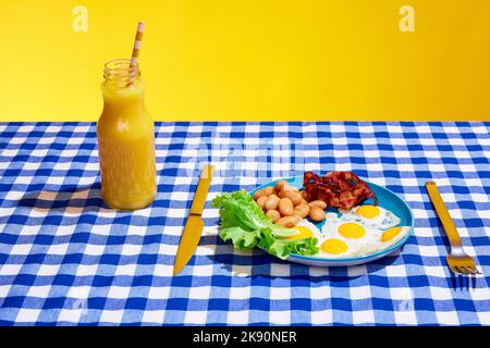 Stillleben. Amerikanisches Frühstück. Spiegeleier mit Speck und Bohnen auf dem Teller ist auf karierten blauen Tischdecken. Interieur im Retro-Stil. Farbenfroh Stockfoto