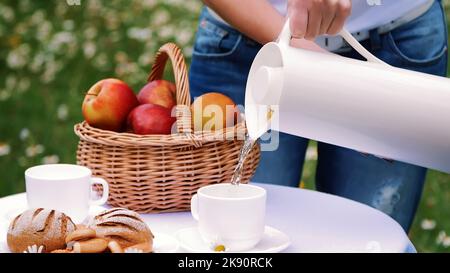Auf dem Hintergrund eines Kamillenrasen gießen Frauenhände Tee aus einem weißen Krug in eine weiße Tasse, eine Thermosflasche. Auf dem Tisch befindet sich auch ein Korb mit roten Äpfeln. Hochwertige Fotos