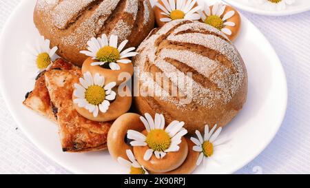 Eine Nahaufnahme, ein weißer Teller mit Gebäck, Keksen und Keksen, verziert mit weißen Gänseblümchen. Auf weißen Untertassen befinden sich zwei weiße Tassen mit Kamillentee. Hochwertige Fotos Stockfoto