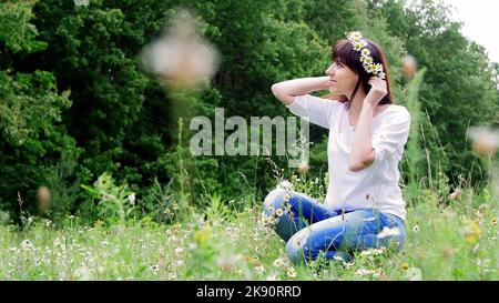 Sommer, inmitten eines Kamillenrasen, in einem Wald, webt eine junge Frau, eine Brünette, einen Kranz aus Kamillen. Hochwertige Fotos Stockfoto