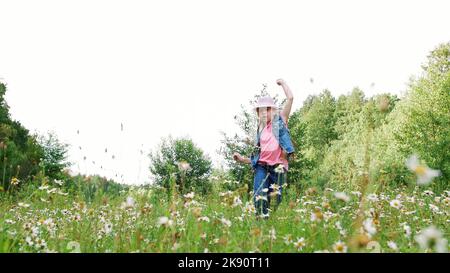 Im Gras, zwischen den Gänseblümchen, auf der Wiese, tanzen, springen, Spaß haben, ein hübsches Mädchen, etwa sieben Jahre alt. Sie hat lange blonde Haare, ist mit einer Jeansweste und einem rosa Hut bekleidet. Hochwertige Fotos Stockfoto