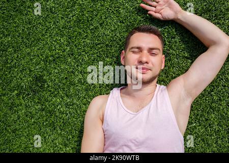 Ein junger Mann in einem rosa T-Shirt liegt an einem sonnigen Tag auf grünem Gras Stockfoto