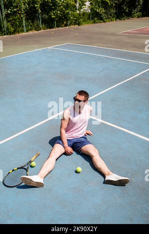 Ein hübscher Mann in einem T-Shirt und Shorts sitzt mit einem Schläger auf einem Tennisplatz. Mann in Sonnenbrille Stockfoto