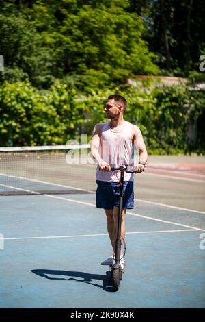 Ein junger Mann mit T-Shirt und Shorts fährt im Park mit einem Elektroroller Stockfoto