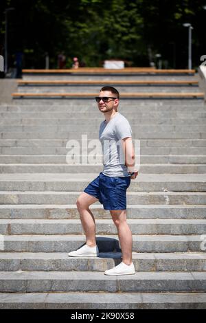 Ein hübscher Mann in einem T-Shirt und Shorts steht auf den Stufen. Mann in Sonnenbrille Stockfoto