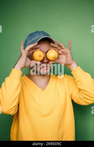 Junger Mann mit einem Bart in einer Mütze auf dem Kopf und Zitronen auf den Augen auf grünem Hintergrund. Porträt eines Mannes in einem gelben Pullover Stockfoto