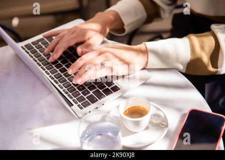 Seitenansicht der weiblichen Hand, die auf der Laptop-Tastatur tippt. Freiberuflicher Mitarbeiter, der im Café mit einem Laptop arbeitet. Technologie und flexibles Arbeiten. Stockfoto