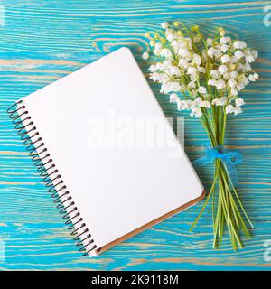 Blumenstrauß Maiglöckchen und leeres Papier-Notizbuch auf blauem rustikalem Tisch von oben, Draufsicht, Platz für Text, flaches Lay. Stockfoto