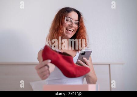 Online-Shopping mit der Anwendung am Telefon. Glückliche Frau packt Schuhe aus und hält ein Smartphone. Die Erwartung und Realität des Kaufs ist Stockfoto