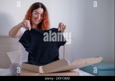 Frau packt online ein T-Shirt aus, während sie im Bett sitzt. Lieferung nach Hause Stockfoto