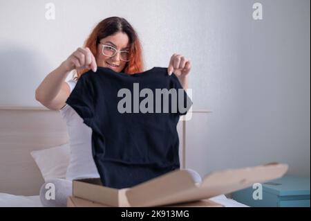 Frau packt online ein T-Shirt aus, während sie im Bett sitzt. Lieferung nach Hause Stockfoto