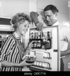 In der Küche 1950s. Ein Paar in der Küche und im Kühlschrank, wo das Bier im Türregal aufbewahrt wird. Fünf Bierflaschen sind zu sehen. Frau hält eine Schüssel mit Garnelen in der Hand. Schweden 1959. Kristoffersson Ref. CH77-1 Stockfoto