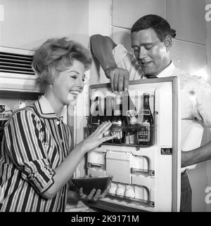 In der Küche 1950s. Ein Paar in der Küche und im Kühlschrank, wo das Bier im Türregal aufbewahrt wird. Fünf Bierflaschen sind zu sehen. Frau hält eine Schüssel mit Garnelen in der Hand. Schweden 1959. Kristoffersson Ref. CH77-3 Stockfoto