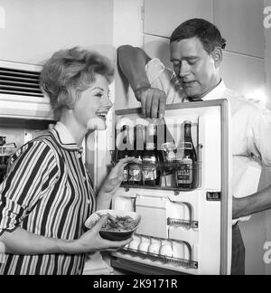 In der Küche 1950s. Ein Paar in der Küche und im Kühlschrank, wo das Bier im Türregal aufbewahrt wird. Fünf Bierflaschen sind zu sehen. Frau hält eine Schüssel mit Garnelen in der Hand. Schweden 1959. Kristoffersson Ref. CH77-2 Stockfoto