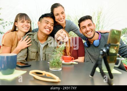 Gruppe glücklicher multirassischer Freunde, die Videoanruf auf dem Smartphone haben, während sie zusammen am Tisch mit Kaffee auf der Terrasse der Cafeteria im Freien sitzen Stockfoto
