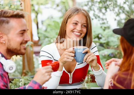 Glückliche junge Frau lächelt und hört Freund beim Kaffee trinken zusammen am Sommertag im Garten Stockfoto