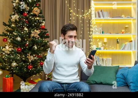 Der Mann, der Sieg und Erfolg feierte, erhielt eine gute Nachricht im Internet, die während der weihnachts- und Neujahr auf dem Sofa in der Nähe des Baumes saß und die Erfolgsgeste per Telefon hielt. Stockfoto