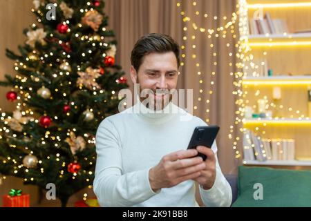 Fröhlicher Mann zu Hause nutzt das Telefon, feiert Weihnachten und Neujahr auf dem Sofa in der Nähe des Weihnachtsbaums, gibt Nachrichten ein und durchsucht online im Internet. Stockfoto