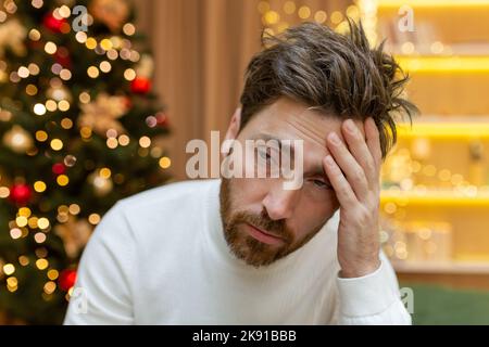 Trauriger, depressiver Mann zu Hause, der auf dem Sofa in der Nähe des Weihnachtsbaums sitzt, Mann, der Weihnachten und Neujahr aus der Nähe feiert. Stockfoto