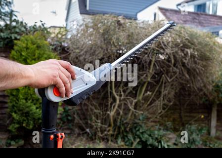 Nahaufnahme von batteriebetriebenen Heckenscheren mit der Hand eines Mannes, der sie vor der Hecke im Garten hält. Stockfoto