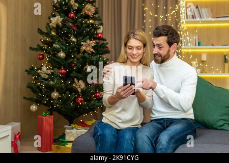 Familienpaar verliebt Mann und Frau zu weihnachten feiern Neujahr sitzen auf dem Sofa zu Hause Video am Telefon und lächeln Auswahl von Geschenken im Online-Shop Einkaufen Remote. Stockfoto