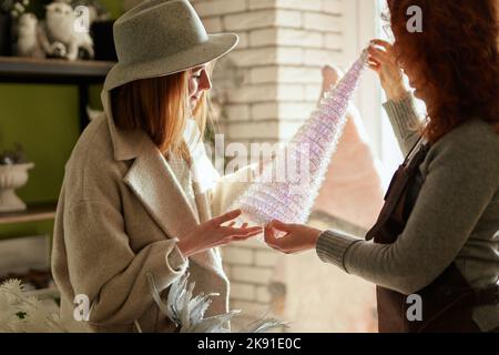 Neujahrsdekoration. Teen shopaholic Mädchen in warmen Kleidern und grauen Hut Auswahl kleiner Weihnachtsbaum im Dekor speichern mit Hilfe von Berater. Winterurlaub Verkaufskonzept. Hochwertige Bilder Stockfoto