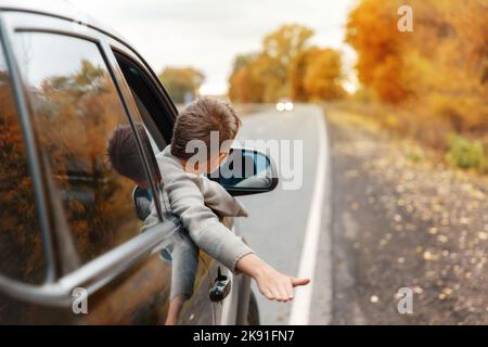 Junge, der seine Köpfe und Hände aus dem Autofenster zieht, während er eine Landstraße hinunter fährt. Bewegungsunschärfe. Kind genießt vom Auto aus den Blick auf die Straße. Fe Stockfoto