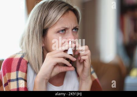 Kranke Frau, die auf dem Sofa sitzt und mit Nasentropfen in eine Decke gehüllt ist Stockfoto