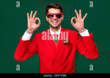 Foto von fröhlich positive santa claus Kerl tragen roten Anzug Brille zeigt zwei okey Zeichen isoliert grünen Hintergrund Stockfoto