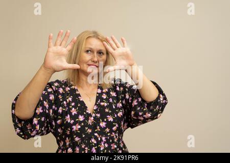 Hübsche blonde Frau mit langen Haaren posiert in dunklem Kleid auf beigem Hintergrund. Emotionales Porträt. Sie macht ein verängstigte Gesicht und hält ihre Hände fest Stockfoto
