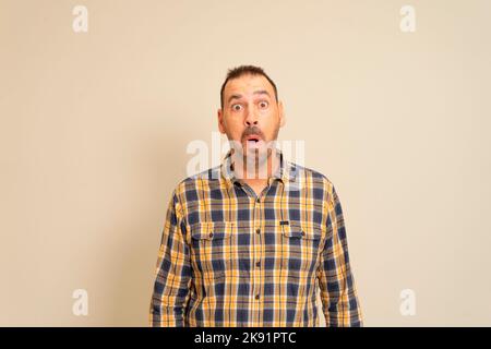Ein hübscher Mann mit kariertem Hemd, der auf einem beigen Hintergrund steht, erschreckt und schockiert von überraschendem Ausdruck, Angst und aufgeregt Stockfoto
