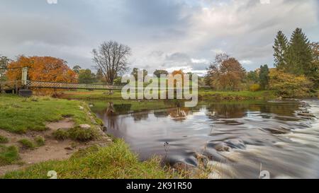 Die Hebden Suspension Bridge erstreckt sich über den Fluss Wharfe in Yorkshire, während sich die Herbstfarben im Wasser spiegeln. Stockfoto