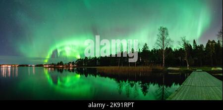 Spektakuläres nächtliches Panorama starke grüne Lichter der tanzenden Aurora über dem nördlichen Wald, Reflexion im See, kleines Boot, Brücke. Stocksjo See Stockfoto