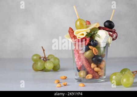 Jarcuterie in einem Glas umfasst verschiedene Arten von Wurst, Gurken, Käse, Nüsse, Gemüse und Obst. Moderne Version von festlichem Snack auf hellgrau Stockfoto