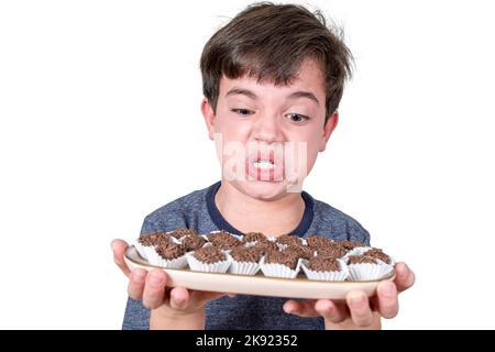 Der 9-jährige Brasilianer hält ein Tablett mit mehreren brasilianischen Fudge-Bällen und macht ein wütendes Gesicht. Stockfoto