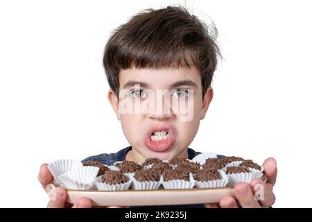 9-jähriges Kind hält ein Tablett mit mehreren brasilianischen Fudge-Kugeln und knirschen seine Zähne. Stockfoto