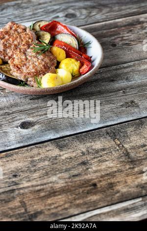Gebratene Koteletts und Salat aus gegrilltem Gemüse Kartoffeln Zucchini roten Pfeffer Rosmarin auf Holzhintergrund mit Textraum. Vertikale Ausrichtung Stockfoto