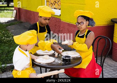 Kinderkochtätigkeit multinationaler Kinder schwarzafrikanischer und kaukasischer Kinder. Fröhliche Gesellschaft von multiethnischen Kindern, die Teig kochen Stockfoto