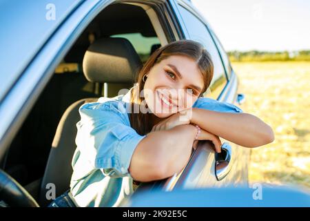 Attraktive junge Frau in blauem Hemd bei Sonnenuntergang mit ihrem Auto Stockfoto