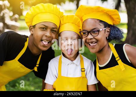 Multinationales Kinderunternehmen kocht in gelben Uniformen lächelnd und grimazing und zeigt Zunge im Freien. Afrikanischer Teenager und schwarzes Mädchen haben Spaß Stockfoto