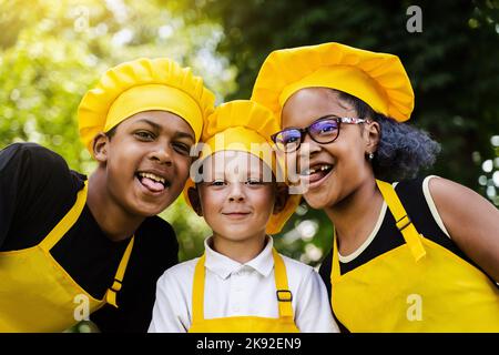 Multinationales Kinderunternehmen kocht in gelben Uniformen lächelnd und grimazing und zeigt Zunge im Freien. Afrikanischer Teenager und schwarzes Mädchen haben Spaß Stockfoto