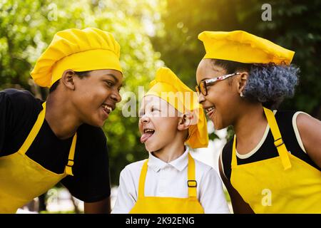 Multinationales Kinderunternehmen kocht in gelben Uniformen lächelnd und grimazing und zeigt Zunge im Freien. Afrikanischer Teenager und schwarzes Mädchen haben Spaß Stockfoto