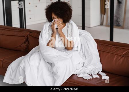 Die kranke afrikanische Mädchengrippe bläst ihr die Nase und niest auf Serviette, während sie zu Hause auf dem Sofa sitzt. Fieber, Kälte und laufende Nase der Frau in Decke gewickelt Stockfoto
