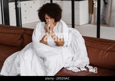Die kranke afrikanische Mädchengrippe bläst ihr die Nase und niest auf Serviette, während sie zu Hause auf dem Sofa sitzt. Fieber, Kälte und laufende Nase der Frau in Decke gewickelt Stockfoto