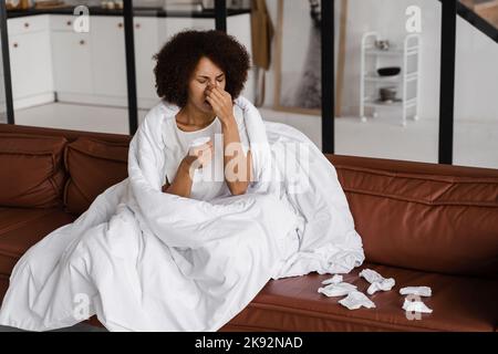 Die kranke afrikanische Mädchengrippe bläst ihr die Nase und niest auf Serviette, während sie zu Hause auf dem Sofa sitzt. Fieber, Kälte und laufende Nase der Frau in Decke gewickelt Stockfoto