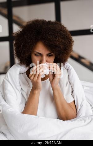 Laufende Nase afrikanische Mädchengrippe bläst ihre Nase und niest in Serviette, während sie zu Hause auf dem Sofa sitzt. Fieber und Erkältung der Frau in Decke gewickelt Stockfoto